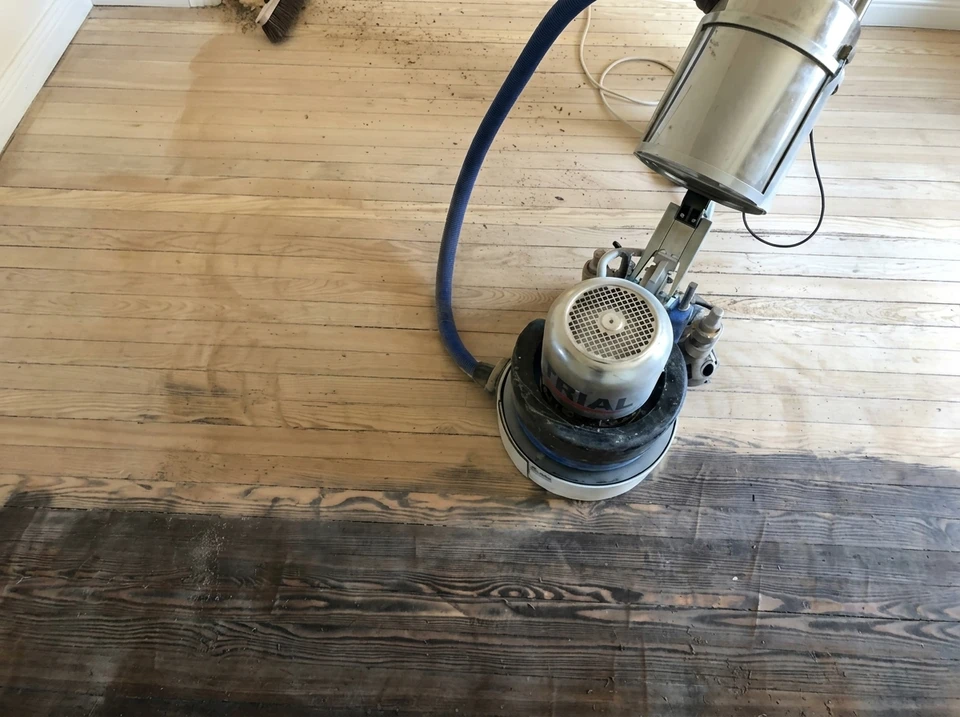 Photographie d'un artisan en tenue professionnelle (chemise et pantalon de travail) effectuant une opération de ponçage ou de vitrification de parquet dans une maison, avec des protections au sol et du mobilier protégé par des bâches, illustrant la propreté du chantier et le sérieux professionnel. L'artisan est concentré sur son travail, utilisant un équipement professionnel performant. L'arrière-plan montre un chantier organisé et propre.